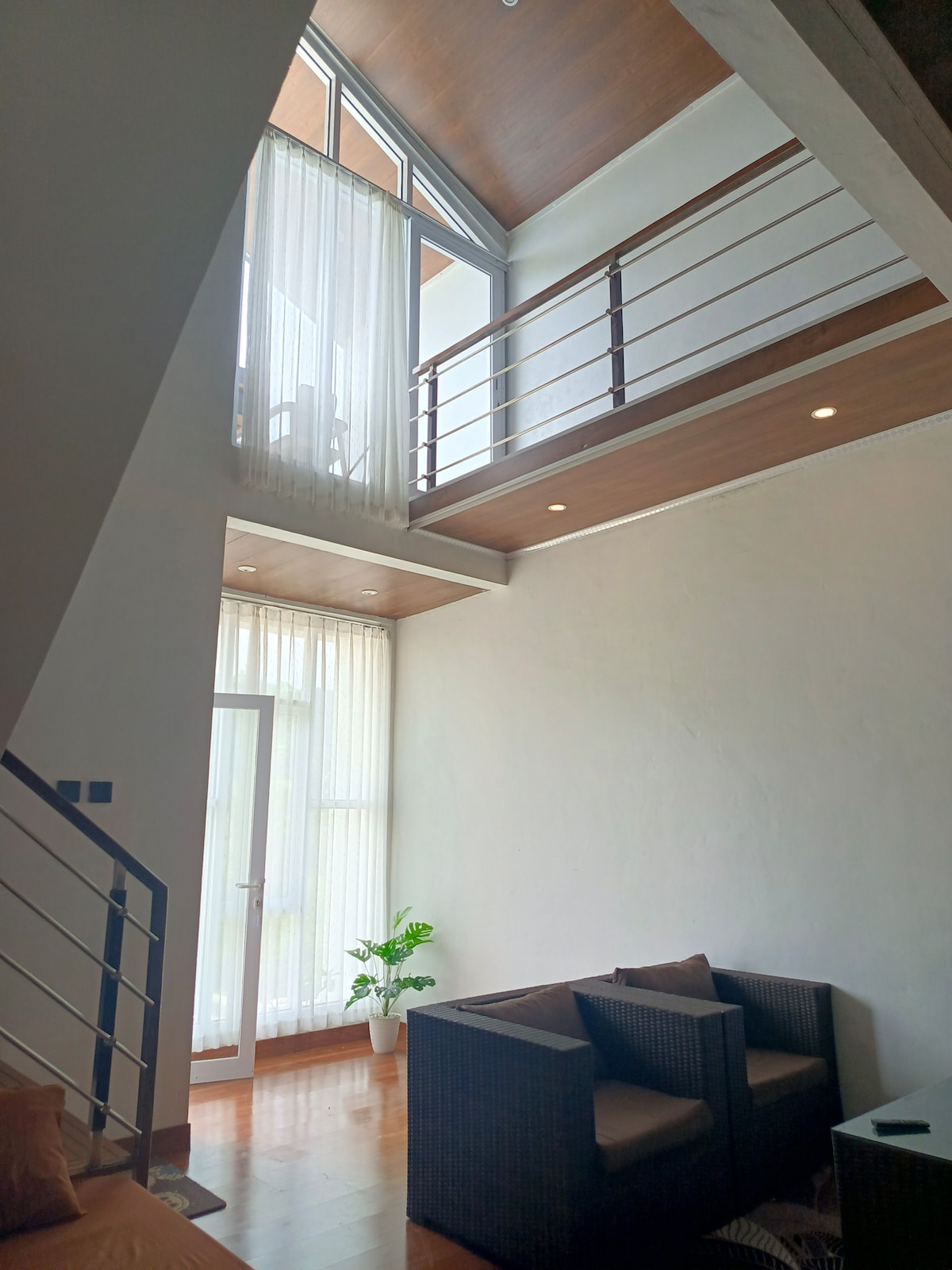 A bright living space is illuminated by natural light filtering through tall, sheer curtains. An open staircase leads to a loft area, while a potted plant adds a touch of greenery. Comfortable seating is arranged for relaxation, enhancing the inviting atmosphere.