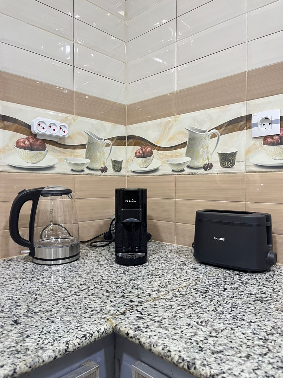 A kitchen countertop is featured, showcasing a sleek black coffee machine, a kettle with a transparent body, and a compact toaster. The design includes decorative wall tiles depicting coffee cups and fruits, enhancing the space's functionality.