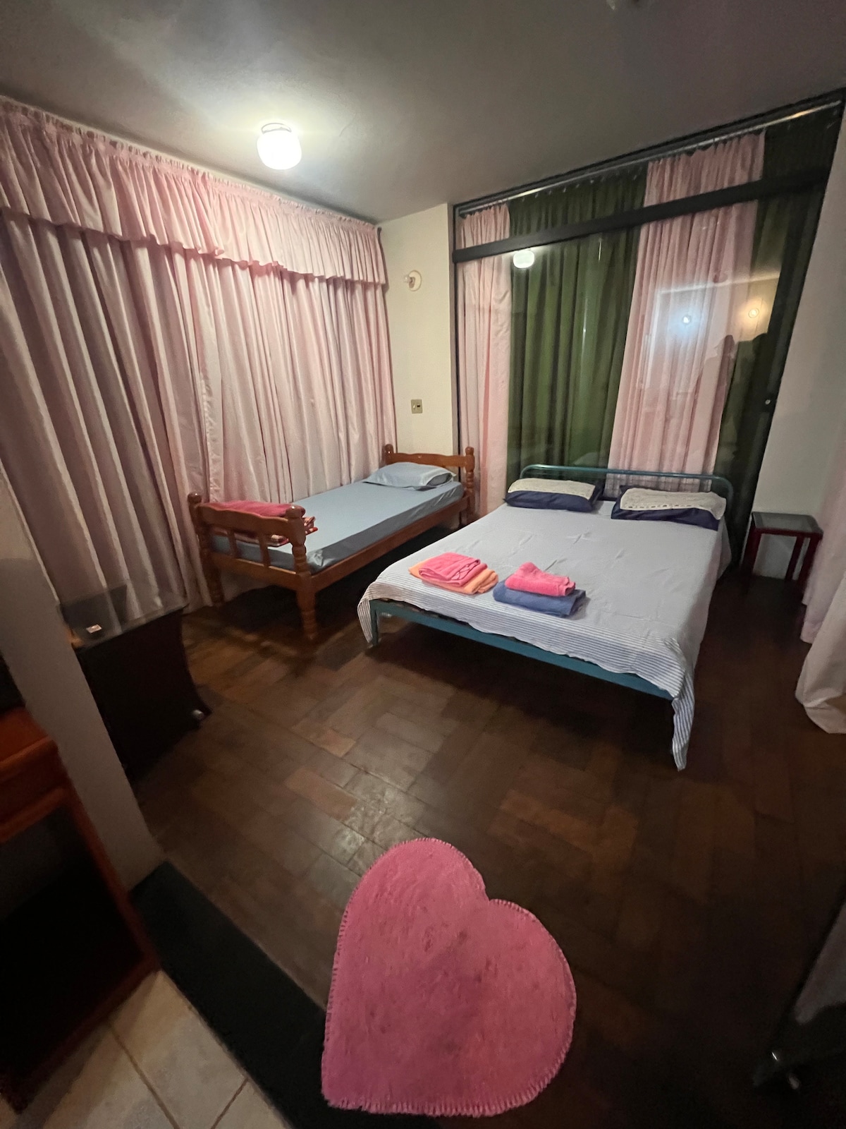 A cozy sleeping area features two beds, one wooden and one metal frame, arranged to allow easy movement. Soft lighting illuminates the room, which has pink and green curtains covering large windows. Fresh towels are neatly placed on one bed, contributing to a welcoming environment.