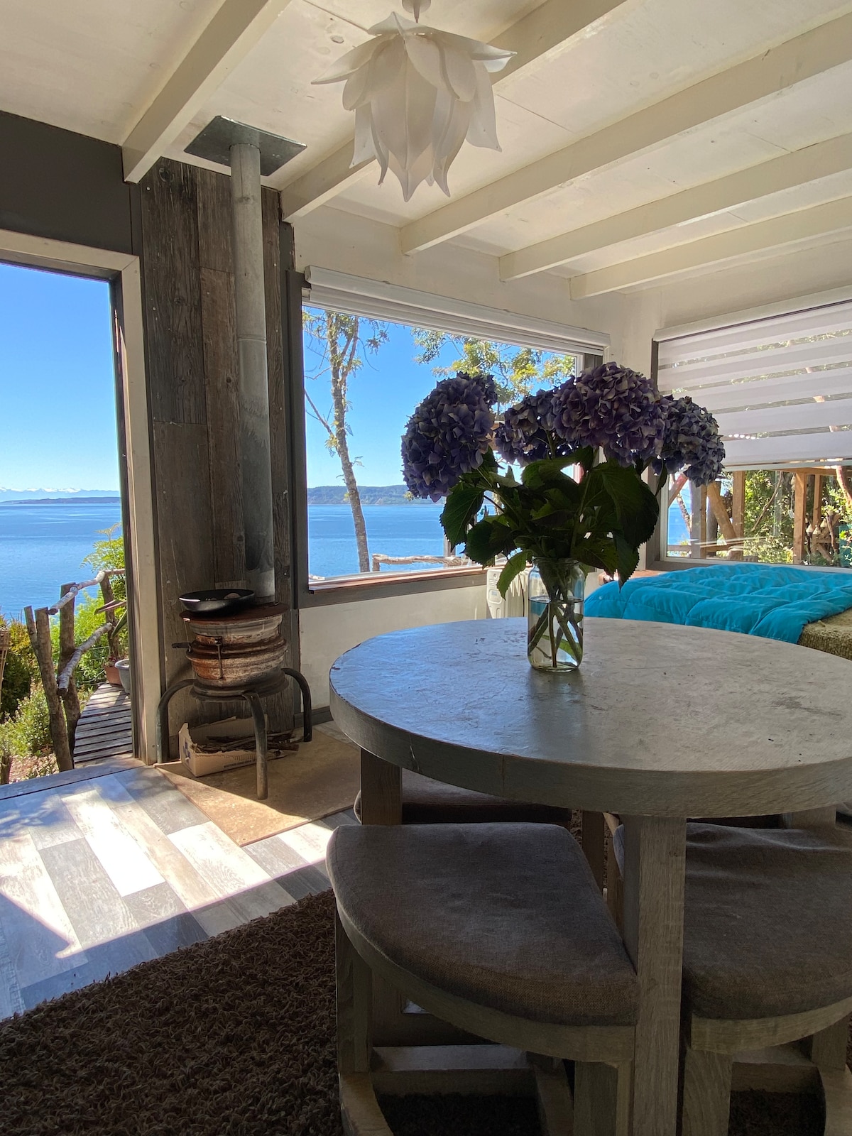 A bright and airy interior features a round dining table with three cushioned chairs, accompanied by a vase of blue hydrangeas. Large windows provide scenic views of the sea and forest, with natural light enhancing the cozy atmosphere.