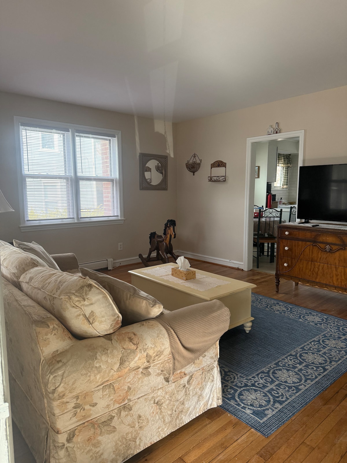 A cozy living area is furnished with a light-colored sofa and a coffee table placed atop a patterned rug. Natural light filters through a nearby window, illuminating the warm wooden floors and decorative accents on the walls.