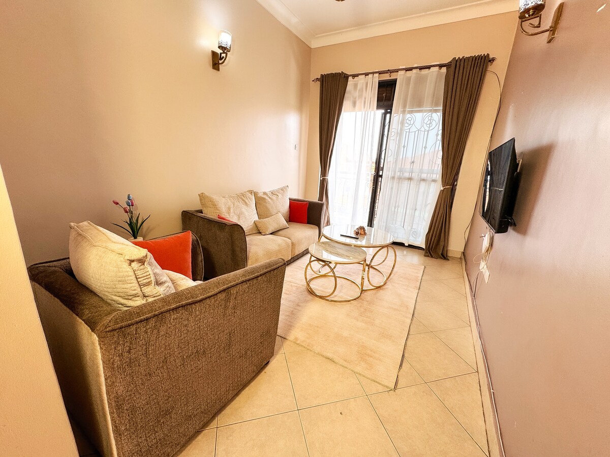 A comfortable living room is highlighted by neutral-toned walls. Two sofas, one brown and one light, are arranged around a round glass coffee table. Natural light filters through sheer curtains covering the balcony door, providing a relaxed atmosphere. A small vase of flowers adds a touch of color.