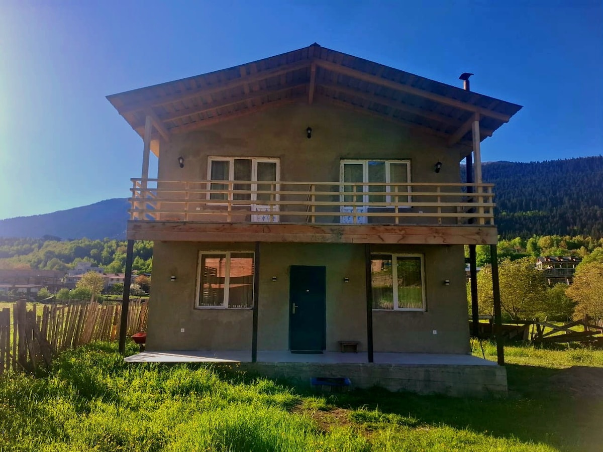 A two-story cottage is presented amidst lush greenery, featuring a wooden balcony on the upper floor. The exterior displays a simple yet functional design with several windows, complemented by natural light and scenic mountain views in the background.