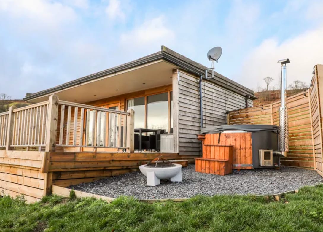 A contemporary lodge exterior is depicted, featuring large glass doors that provide easy access to a wooden deck. A rustic hot tub and a fire pit are visible on the stone pavement, while the surrounding area is complemented by green grass and a sky with scattered clouds.