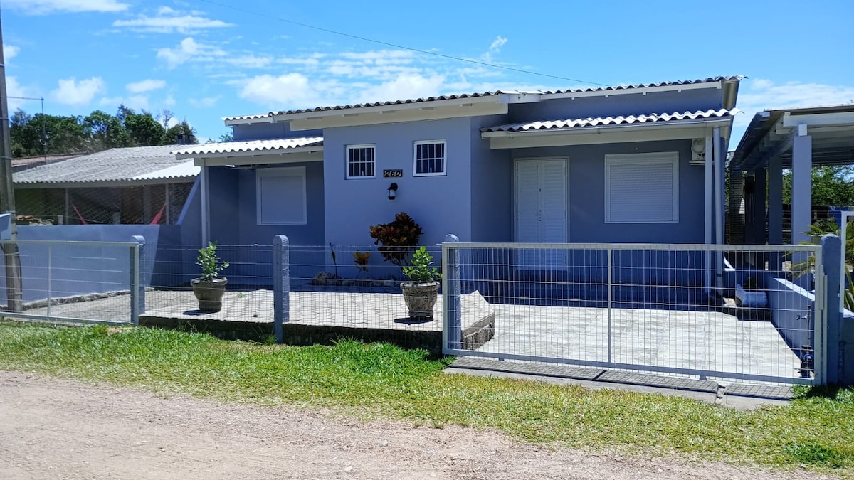 The exterior of the house features a light blue facade with a pitched roof. A neat gray pathway leads to the entrance, surrounded by small planters. A fenced area borders the property, enhancing security and privacy, while a bright sky adds to the overall setting.