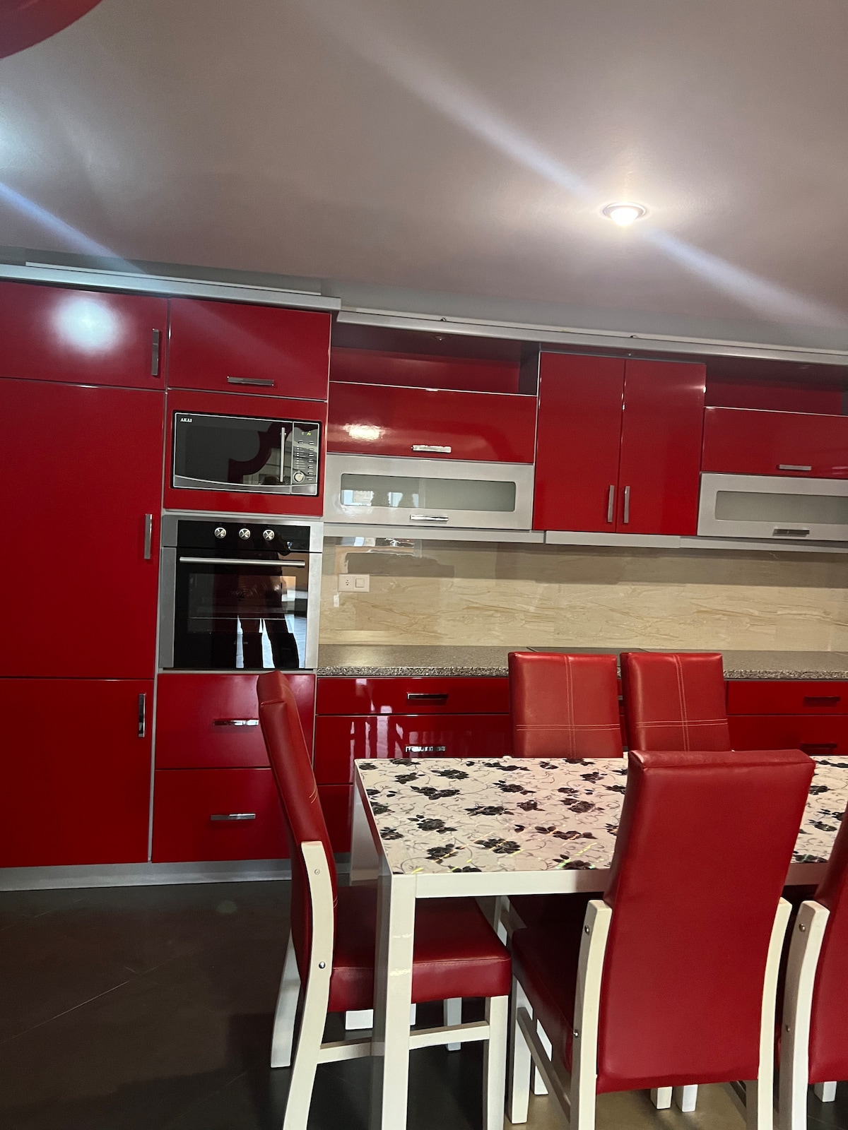 A modern kitchen is highlighted by vibrant red cabinetry and sleek appliances. A glossy table is surrounded by six matching chairs. The light-colored backsplash contrasts with the cabinetry, while overhead lighting provides illumination throughout the space.