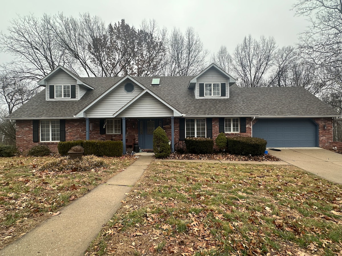 A spacious two-story home with a brick facade is shown on a cloudy day. The front porch is flanked by neatly trimmed shrubs. A driveway leads to a garage on the right, and fallen leaves cover the lawn, adding a natural touch.