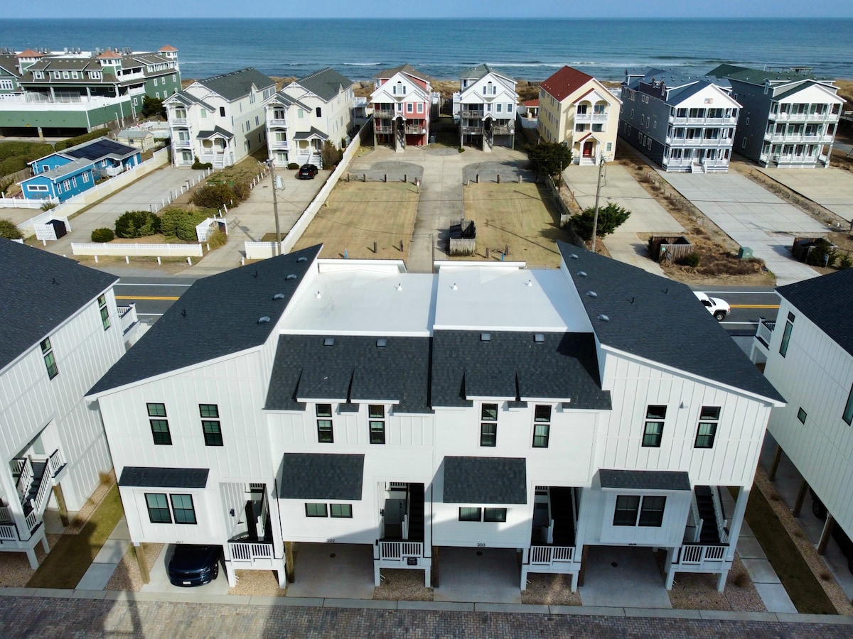 An aerial view captures the modern townhome alongside a stretch of sandy beach. The building features a clean white exterior, with multiple balconies visible. The coastline and ocean are seen in the background, while neighboring houses and a grassy area are also present, emphasizing the community setting.
