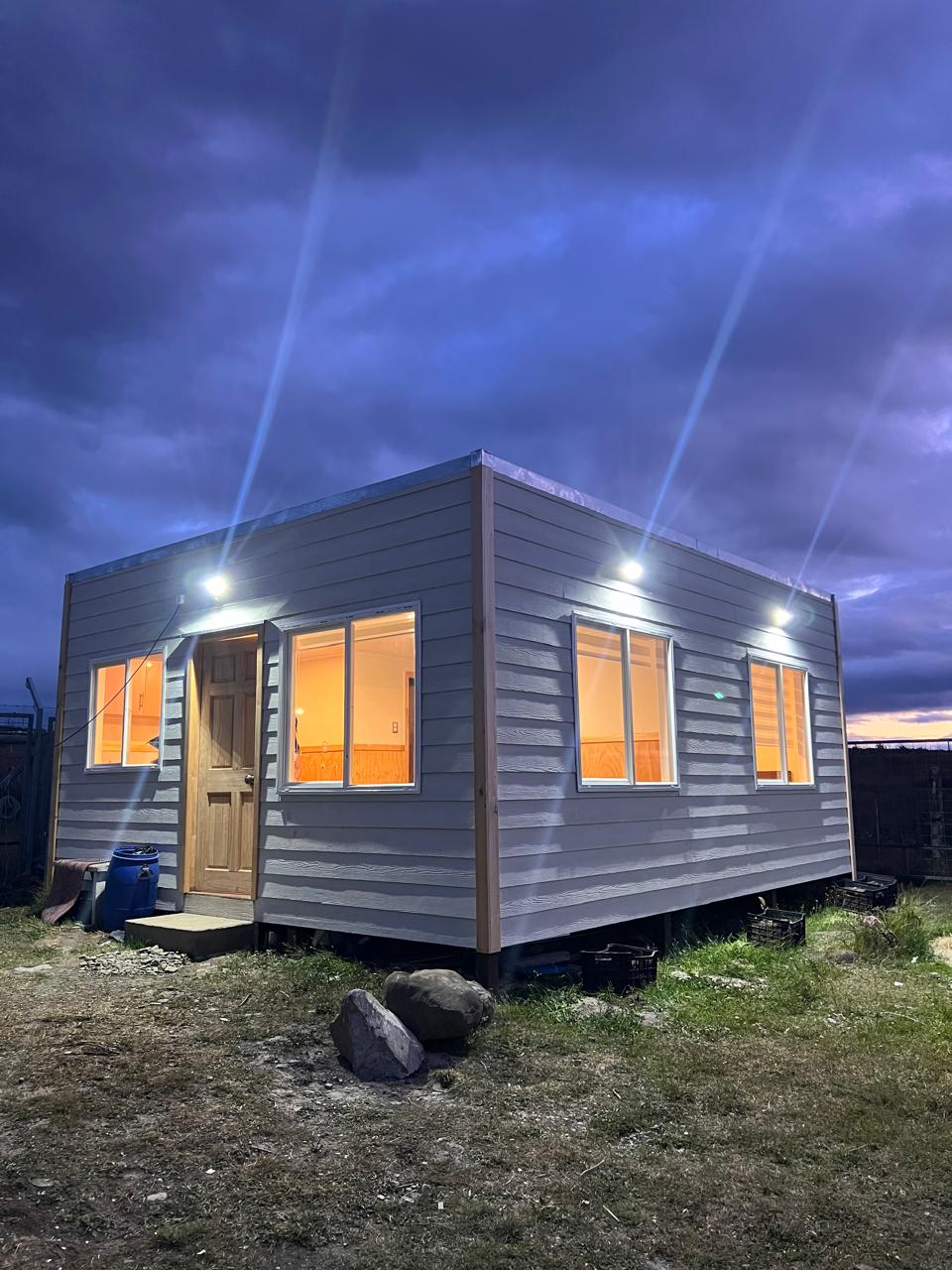 A newly built cabin is shown at dusk, with warm lights glowing from large windows. The structure features a wooden door framed by multiple windows, illuminating the grassy area surrounding it. Outdoor lighting enhances visibility along the exterior.