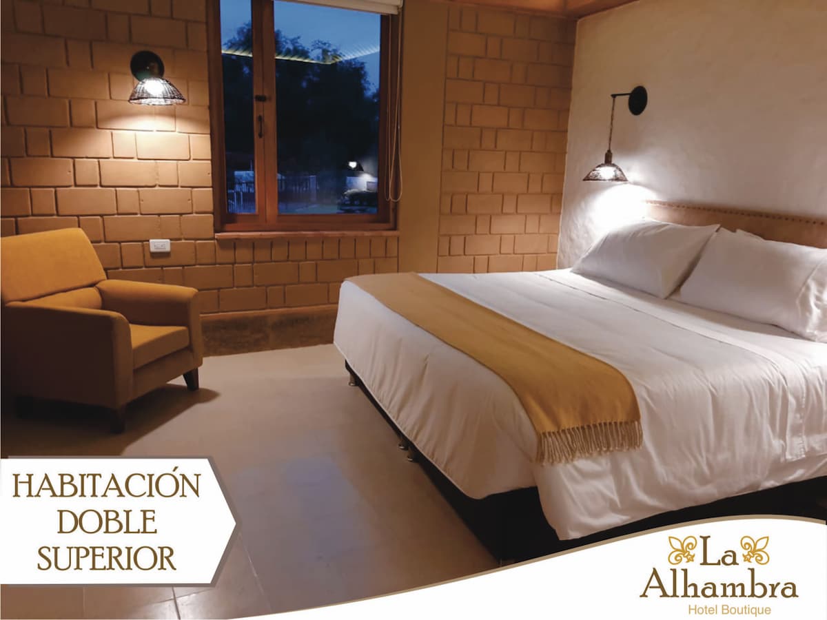 A spacious guest room features a large bed with crisp white linens and a soft throw blanket. A comfortable armchair is positioned near a window that allows natural light to brighten the room, complemented by modern wall sconces.