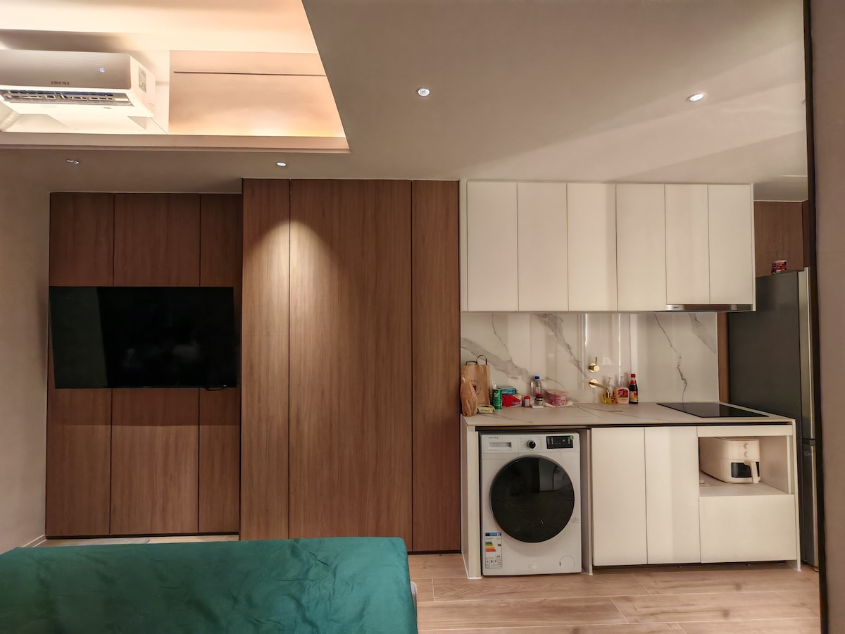 A compact living area features warm wooden cabinetry paired with sleek white kitchen units. A washing machine is positioned next to a simple countertop that includes essential kitchenware. Soft lighting illuminates the space, creating a functional and welcoming environment.