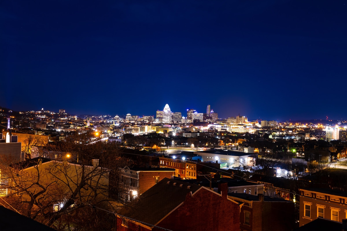 The skyline of Cincinnati is captured at night, displaying illuminated buildings and vibrant city lights against a clear, dark sky. Rooftops and trees are visible in the foreground, creating depth to the view of the bustling cityscape.