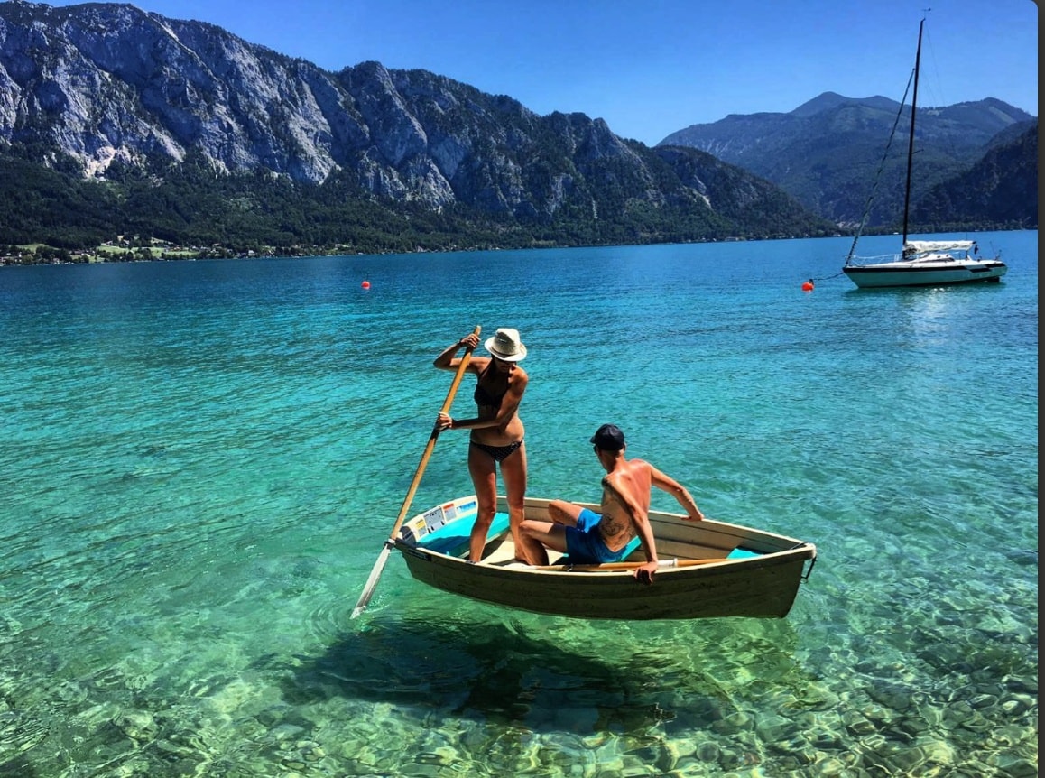 Two individuals are seen in a small wooden boat, gently gliding across clear turquoise waters. The backdrop features majestic mountains under a clear blue sky. A larger sailboat is anchored nearby, completing the serene lakeside scene.