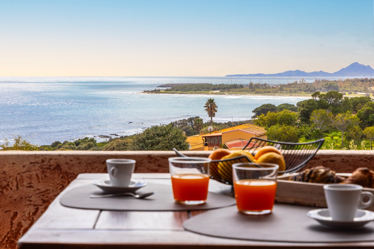A serene view from a balcony is captured, showcasing a coastal landscape with clear blue waters and distant mountains. A table is set with cups of coffee and glasses of orange juice, complemented by a bowl of fresh fruit, inviting relaxation and enjoyment of the surroundings.