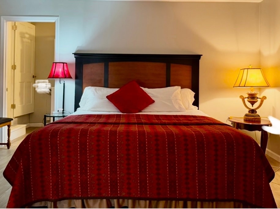 A spacious bedroom features a large bed covered with a red patterned blanket. Soft lighting is provided by a bedside lamp and a decorative table lamp. White linens are visible, creating a comfortable setting. A doorway leads to another room in the background.