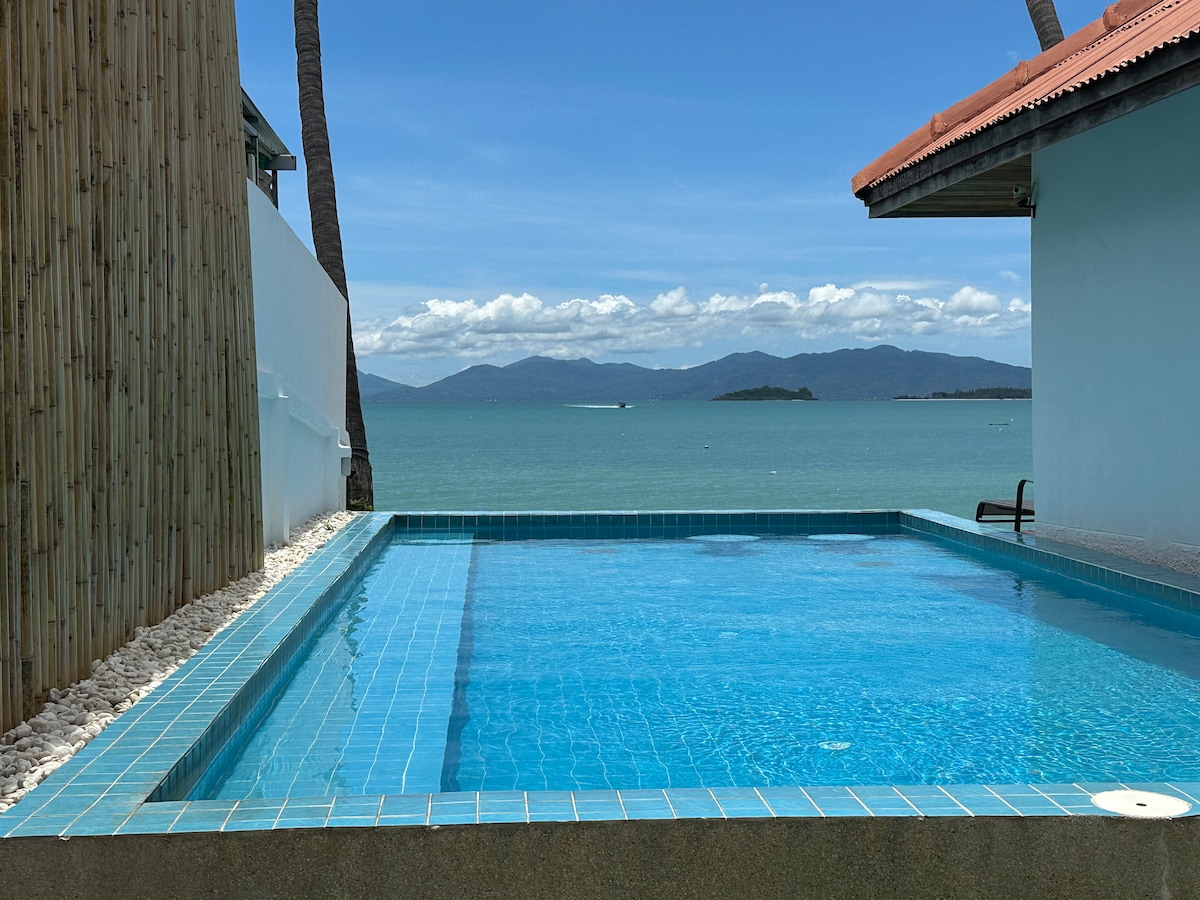 An outdoor swimming pool is framed by a smooth stone border, with clear blue water reflecting the sky. Lush greenery and palm trees provide a natural backdrop, while distant mountains are visible across the calm sea in the background.
