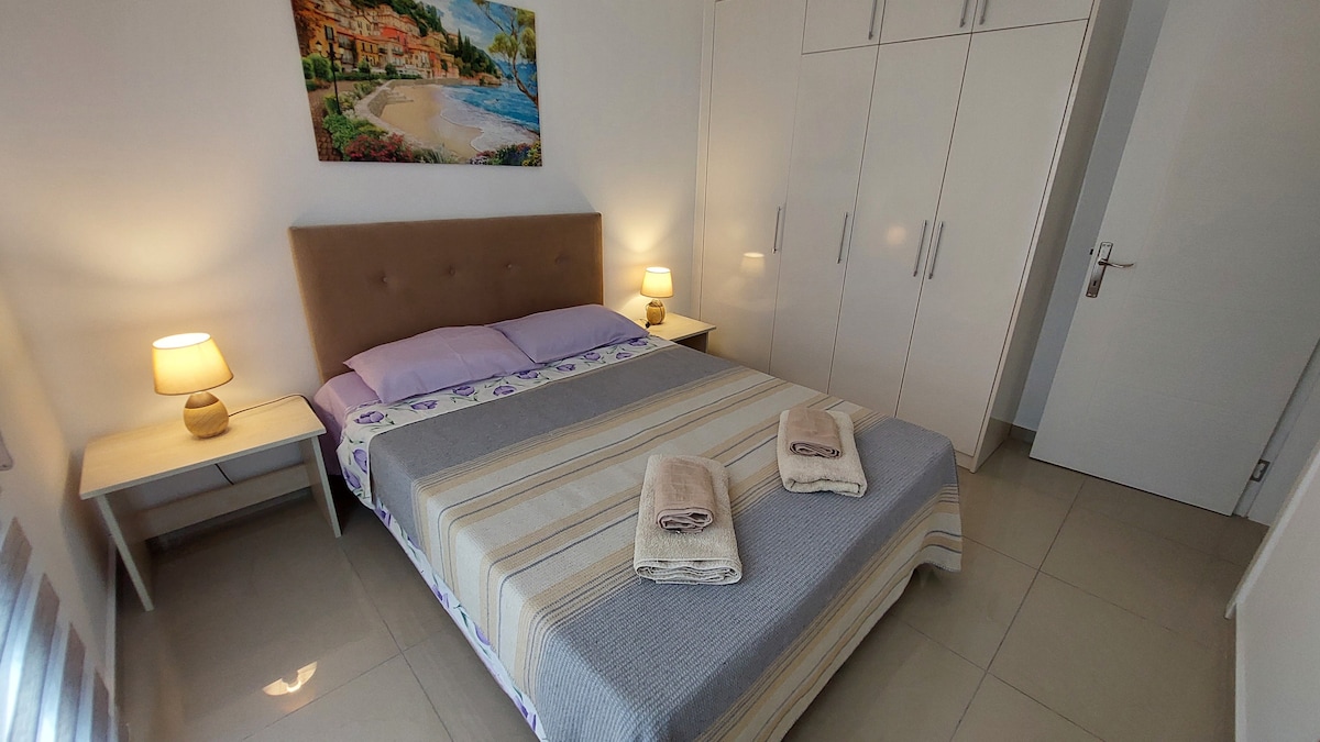 A well-appointed bedroom displays a spacious bed with a light purple blanket and decorative pillows. Two bedside tables with lamps provide soft lighting. A painting of a coastal scene adorns the wall above. Wardrobe doors reflect a clean, modern design.