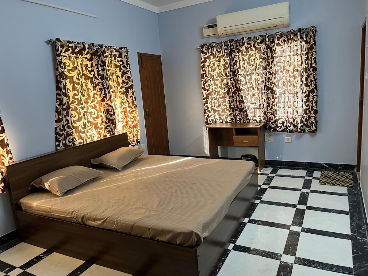 A neatly arranged bedroom features a spacious bed with light bedding. Natural light filters through patterned curtains, enhancing the room's ambiance. A work station with a chair is positioned against the wall, and the floor showcases a classic black and white tile pattern.