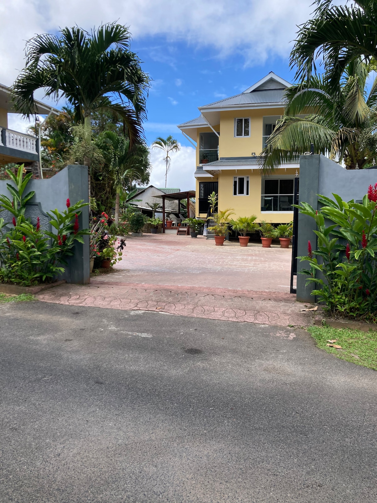 A welcoming entrance is framed by lush green plants and colorful flowers. The driveway leads to the chalet, where a two-story building stands surrounded by palm trees. A shaded area with seating is visible to the side, inviting relaxation.