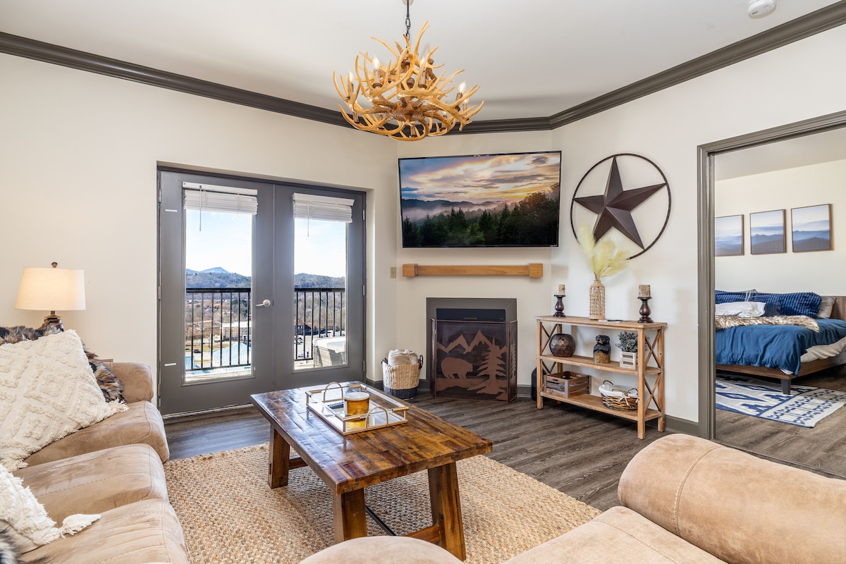 A comfortable living area is showcased with a large sectional sofa and a rustic wooden coffee table. A mounted TV displays a scenic mountain view. Large doors lead to a balcony, while a decorative star and wall art enhance the inviting ambiance.