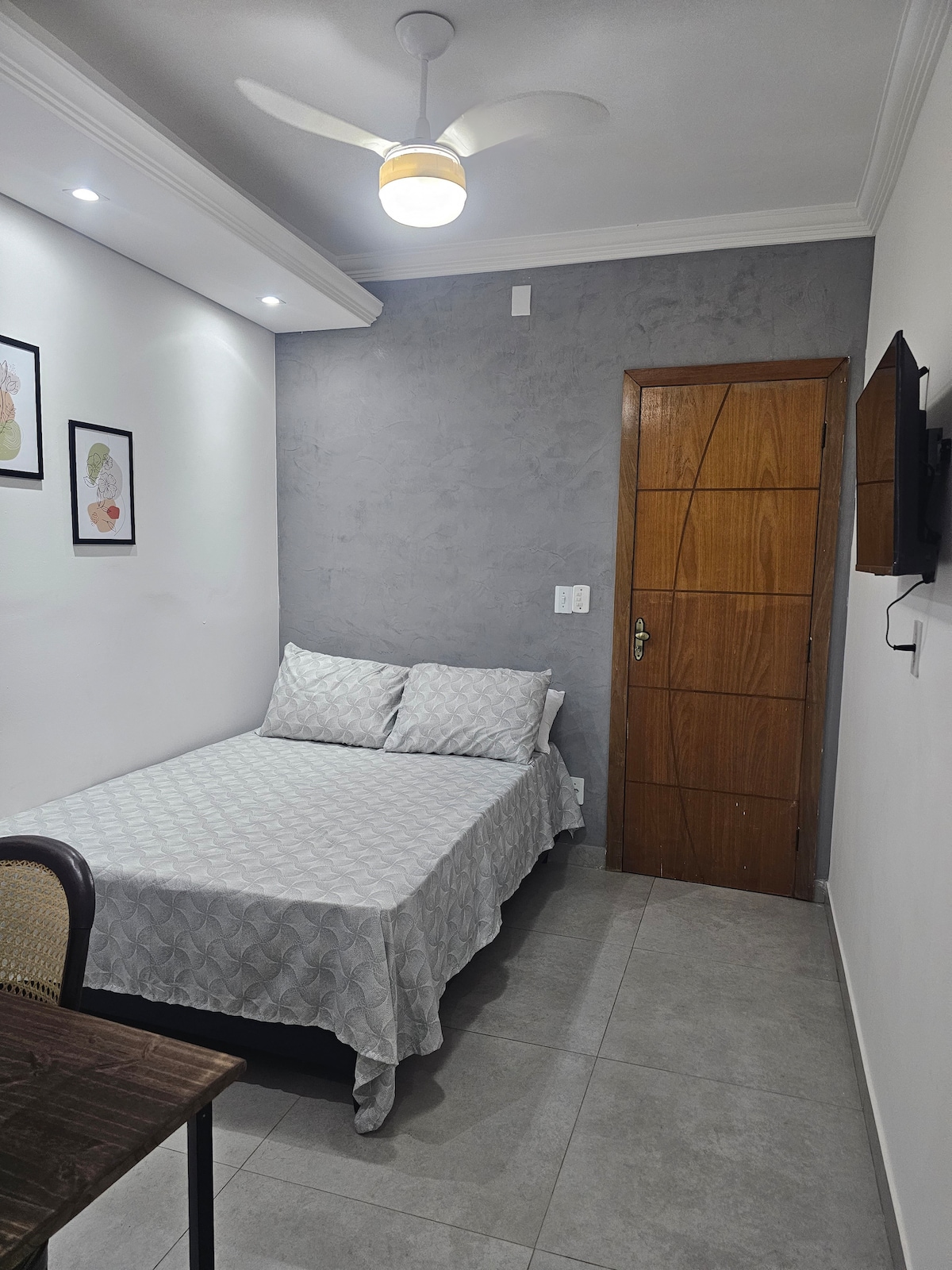 A cozy sleeping area features a spacious bed adorned with light grey bedding. A wooden door and a mounted TV are visible, while artwork is displayed on the walls. Soft lighting is provided by ceiling fixtures, creating a calm atmosphere.