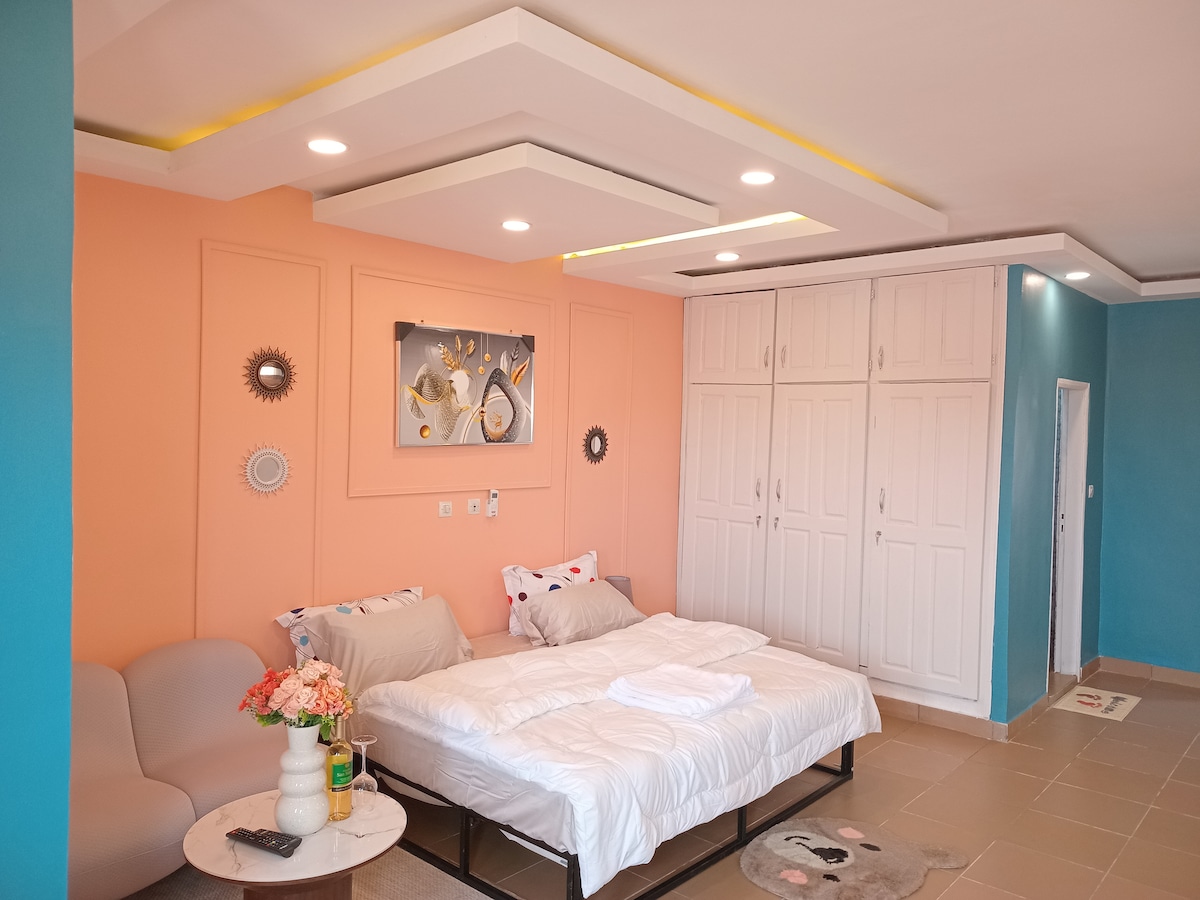 A spacious sleeping area features a large, soft bed with neutral linens against a peach-accented wall. Flanking the bed are a comfortable gray sofa and a small round table with a floral arrangement. Built-in cabinets offer ample storage, and light fixtures illuminate the room.