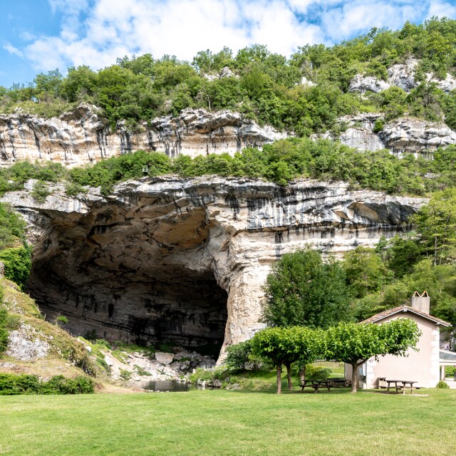 A large cave entrance is framed by lush greenery and rocky cliffs, creating a natural shelter. In the foreground, a grassy area includes a small pink building and several trees, providing a peaceful setting for relaxation and exploration.
