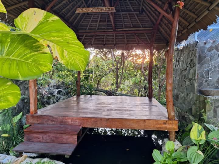 A serene outdoor pavilion is set on wooden flooring, surrounded by lush greenery. Stone walls provide a natural backdrop, while soft sunlight filters through the trees, creating a tranquil ambiance. Steps lead up to the pavilion, inviting relaxation in a peaceful environment.