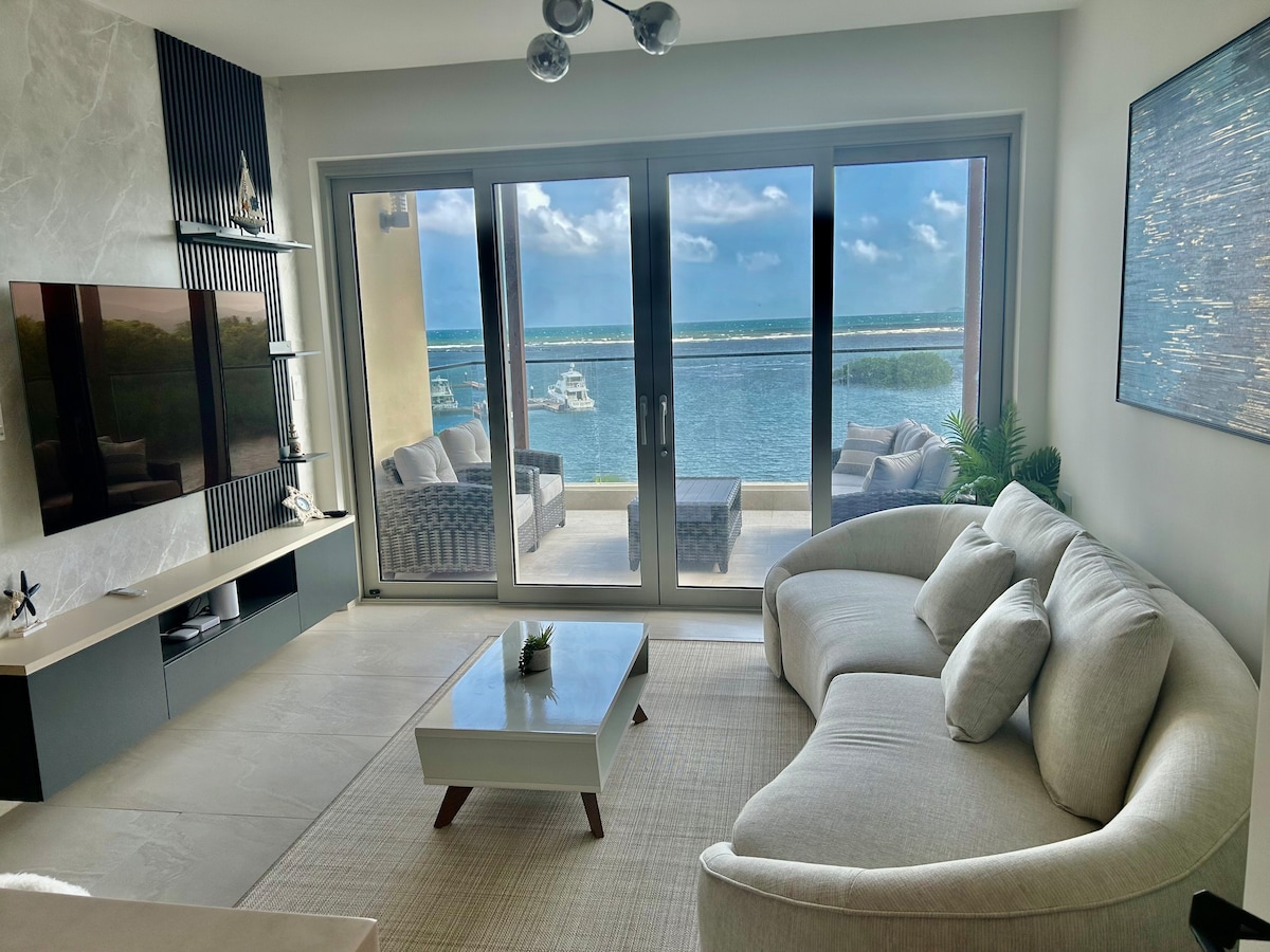 A bright, open living area features large sliding glass doors that provide a view of the water. A light-colored sectional sofa is positioned next to a modern coffee table, and a sleek television is mounted on the wall. Natural light fills the room, creating a relaxed atmosphere.