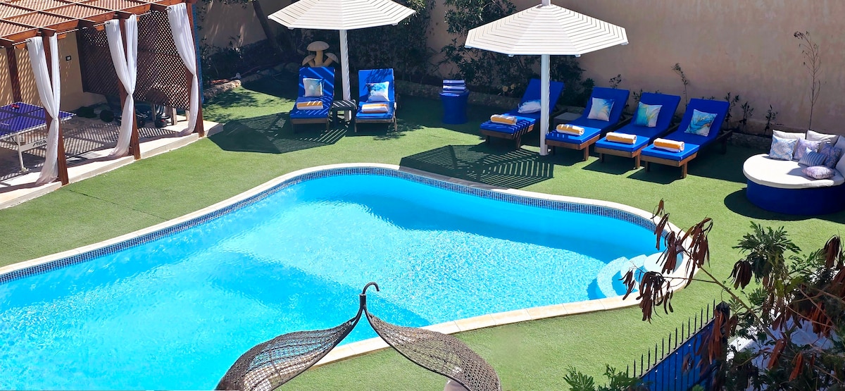 The outdoor area features a shimmering swimming pool surrounded by well-maintained grass. Several sun loungers with blue cushions offer a comfortable spot to relax. Two large umbrellas provide shade, while a circular seating space invites guests to unwind by the poolside.