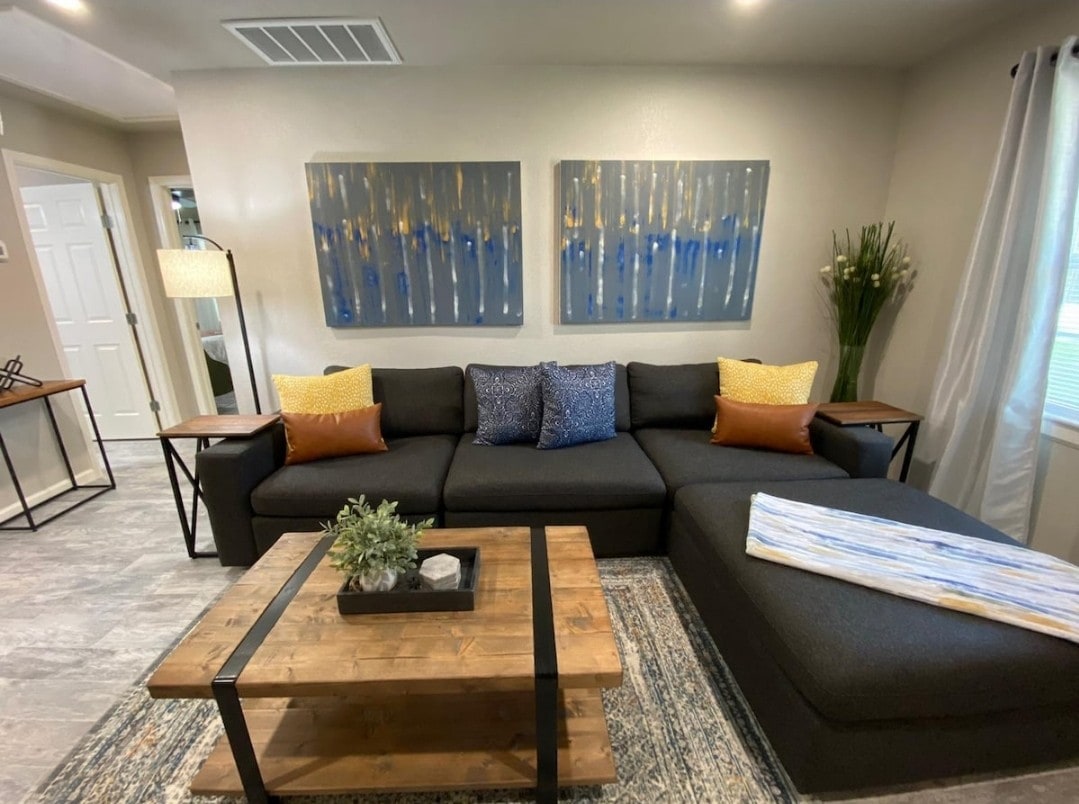A comfortable living area is presented with a spacious dark sofa adorned with colorful throw pillows. A wooden coffee table with a small plant centerpiece sits on a patterned rug. Two large wall art pieces have a soothing blue and silver colour scheme, complementing the light decor.