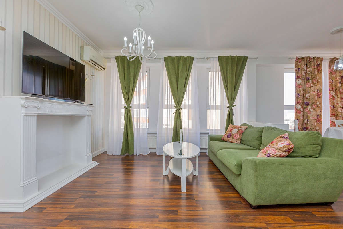 A living area is presented with wooden flooring and large windows covered by sheer curtains. A green sofa adorned with decorative cushions forms a seating area, complemented by a round coffee table. A chandelier hangs from the ceiling, adding a touch of elegance to the space.