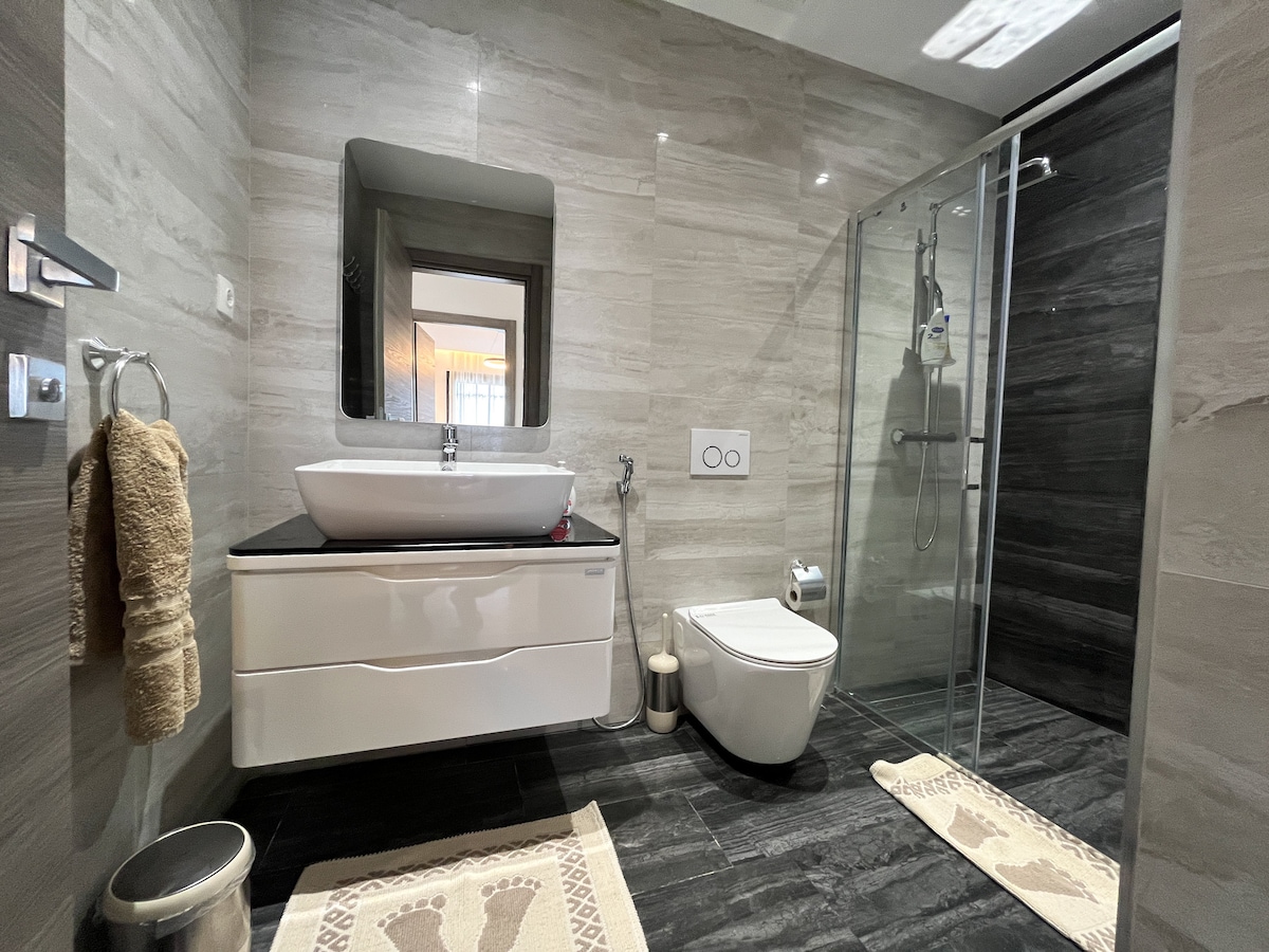 The spacious bathroom is equipped with modern fixtures, featuring a large sink atop a sleek vanity. A glass shower stall is visible, alongside a contemporary toilet. Soft towels are neatly arranged, and textured bath mats add a touch of comfort to the stylish floor.
