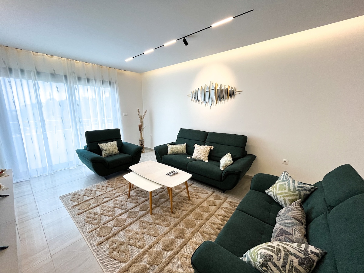 A spacious living area features two dark green sofas and a lighter green coffee table. Soft, patterned cushions add comfort to the seating. Natural light filters through sheer curtains covering glass doors, illuminating the light tile flooring and enhancing the minimalist decor.