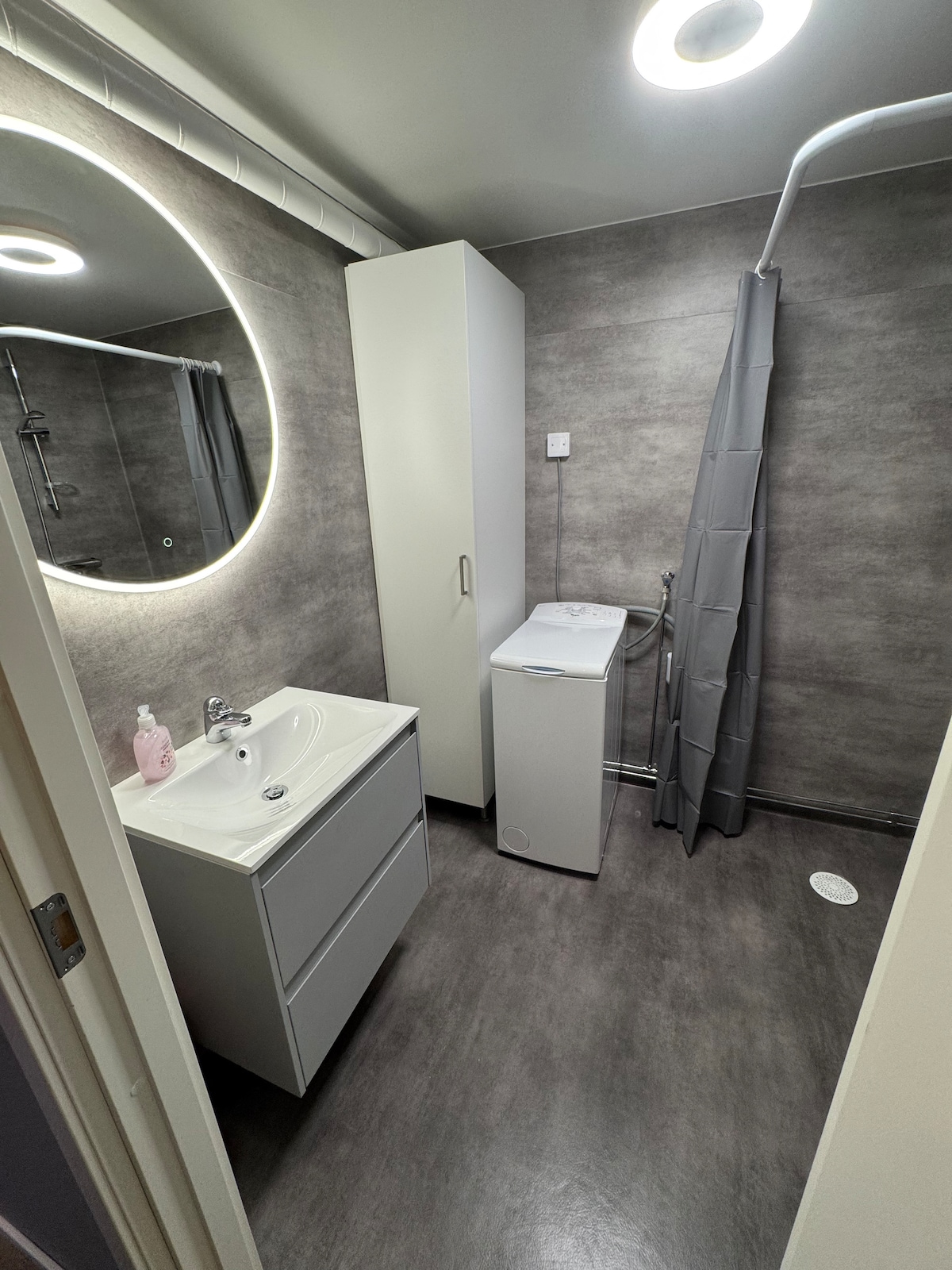 A modern bathroom is featured, equipped with a compact sink and a storage cabinet. A circular mirror reflects soft, indirect lighting. An enclosed shower area with a light grey curtain and a washing machine are visible, highlighting functionality in a sleek design.