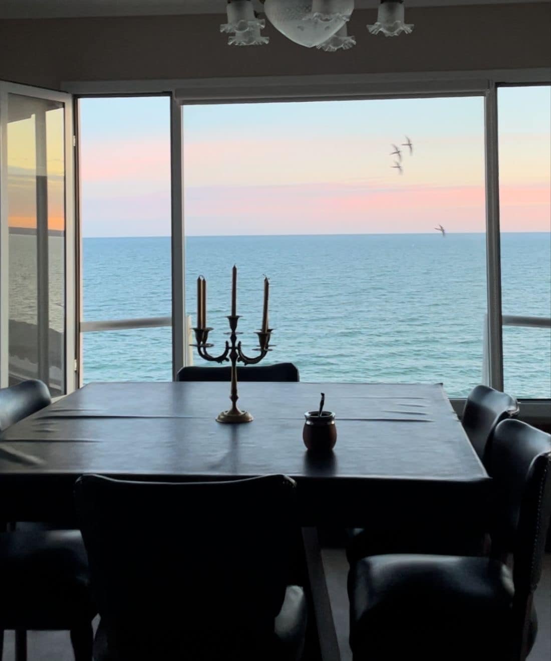 A dining area is depicted with a dark table at its center, surrounded by black chairs. Large glass doors provide panoramic views of the ocean, with a soft gradient of colors in the sky. A candle holder sits on the table, adding to the tranquil setting.