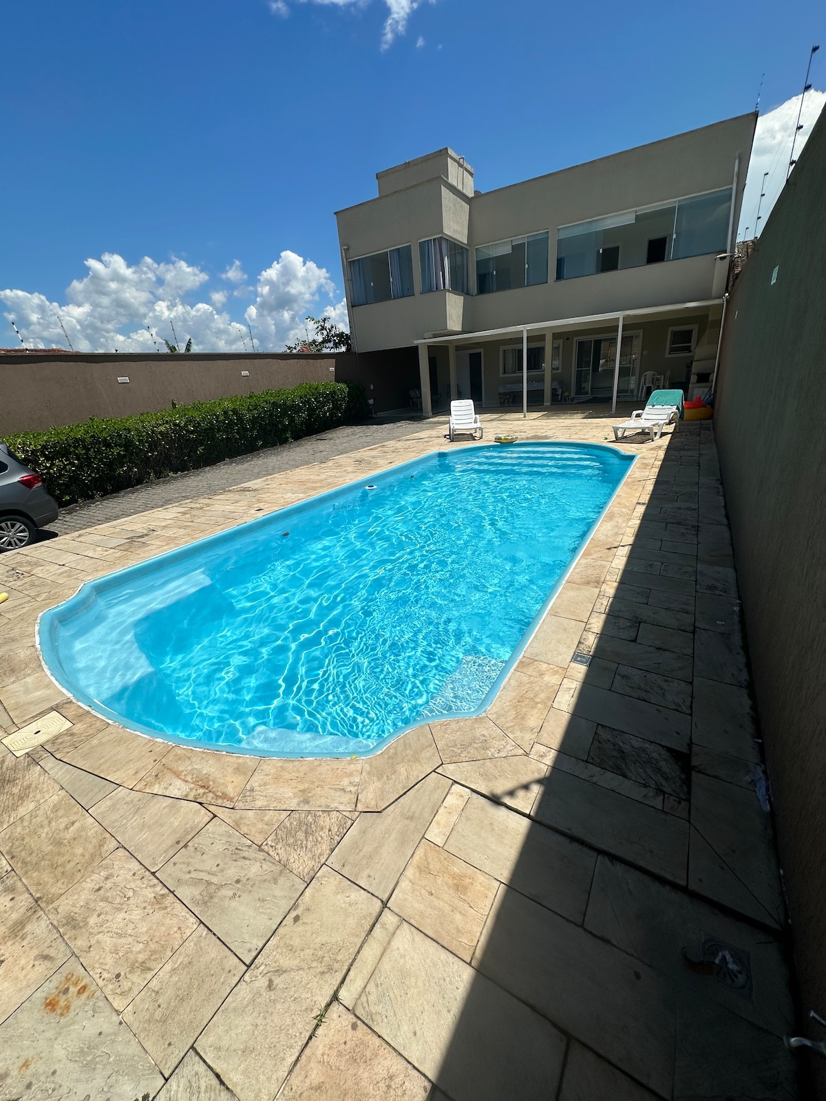 A rectangular swimming pool is surrounded by a spacious patio, featuring natural stone tiles. Sun loungers are positioned along the pool's edge. The two-story house in the background has large glass windows, allowing for ample natural light. Lush greenery borders the property.