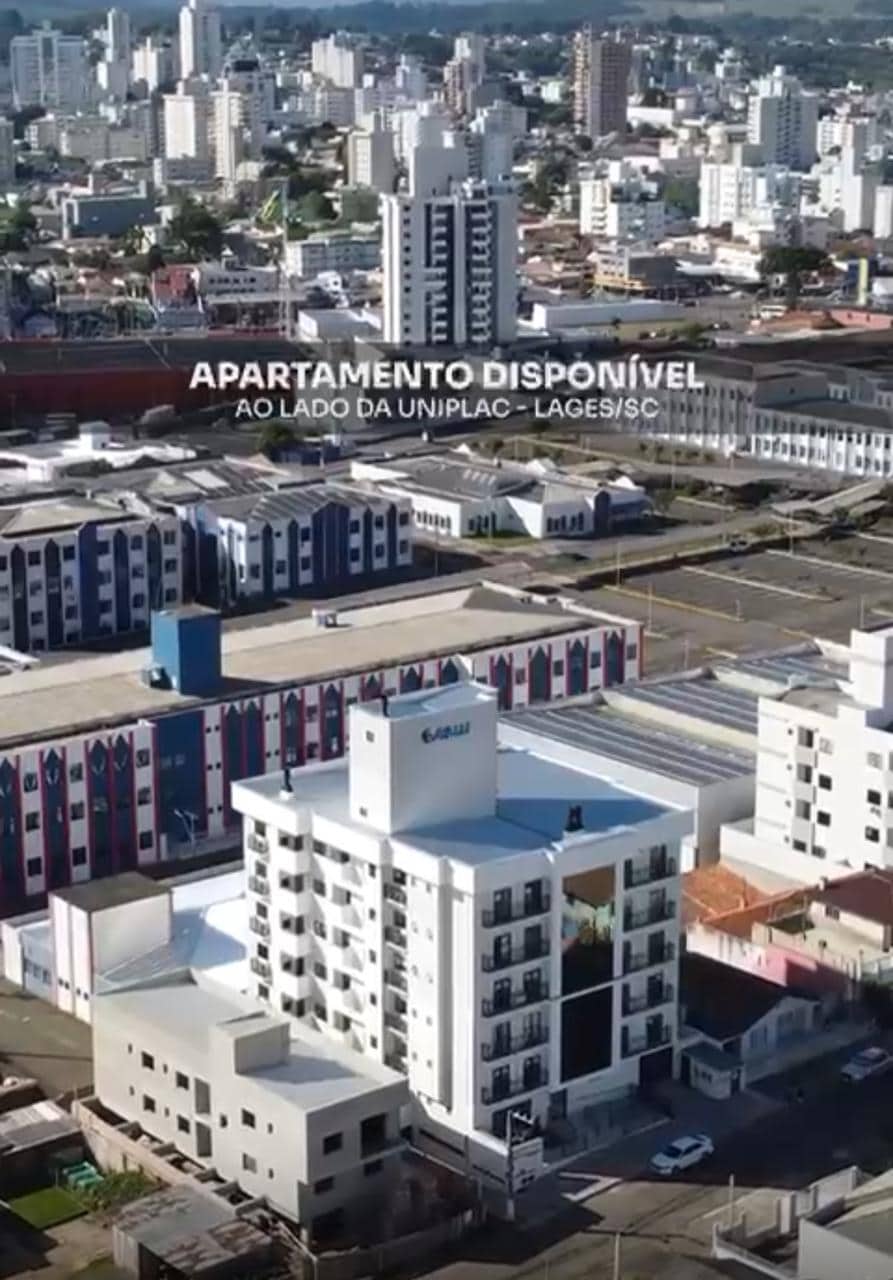 An aerial view showcases a multi-story building situated near the Uniplac campus in Lages, SC. The surrounding urban landscape includes a mix of tall buildings and low-rise structures, with streets and greenery visible in the distance.