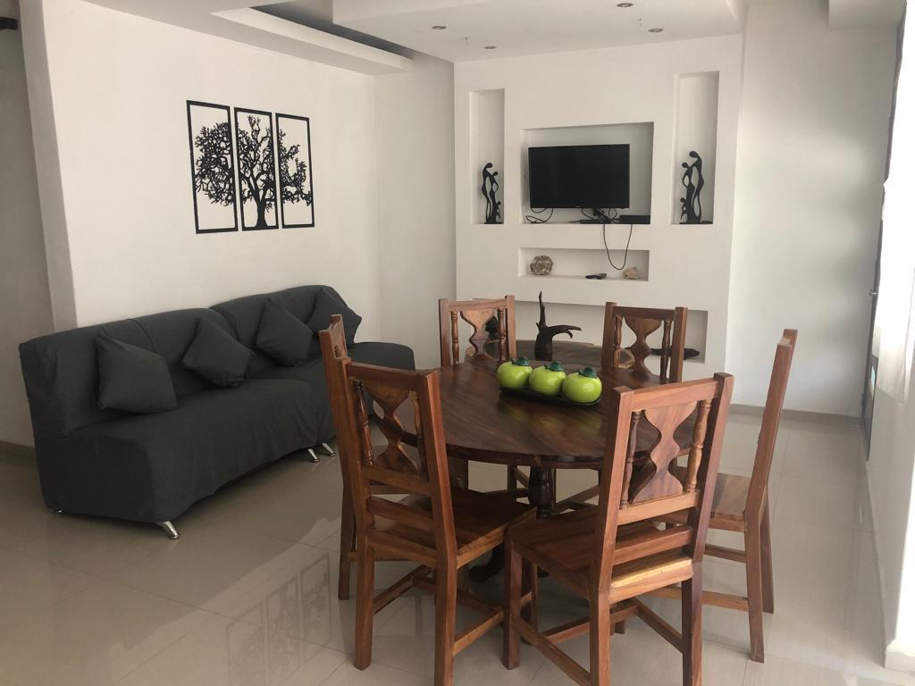 A living area features a gray sofa equipped with decorative cushions, positioned adjacent to a round wooden dining table surrounded by six chairs. A television is mounted on the wall above a minimalist shelf, complemented by artwork and decorative elements.