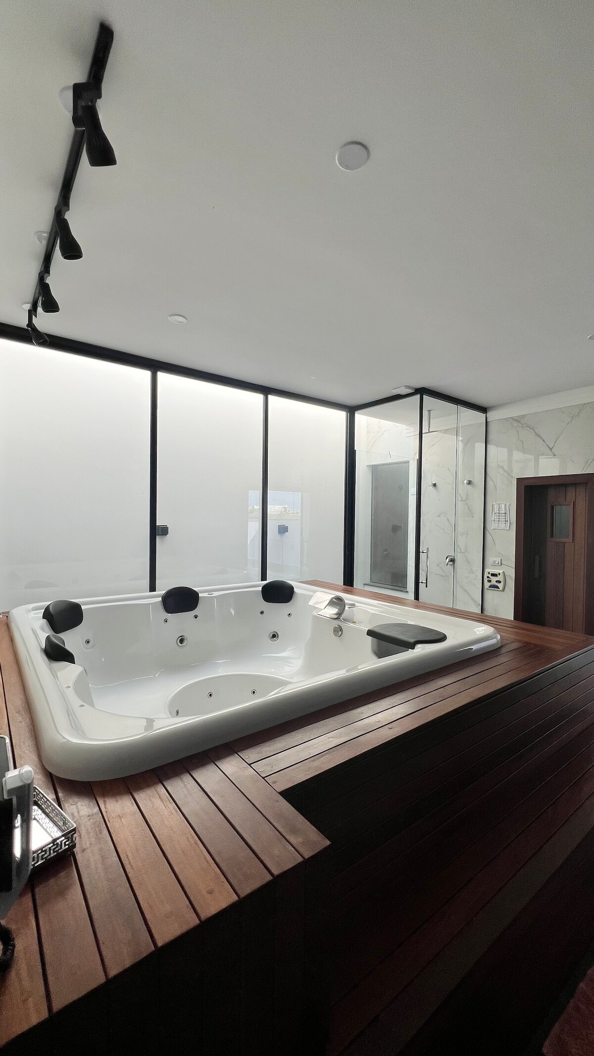 A modern jacuzzi is positioned on a wooden deck, within a light-filled room featuring floor-to-ceiling windows. The design includes sleek, minimalistic lines, with a glass shower enclosure visible in the background, creating an elegant and relaxing setting.