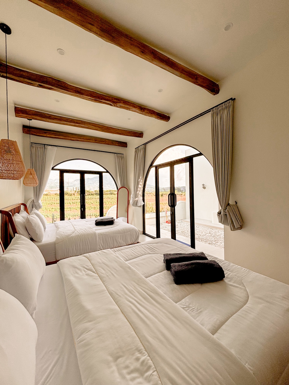A light-filled bedroom features two plush beds, each adorned with inviting bedding. Large glass doors provide access to the outdoor area and frame views of the surrounding greenery. Natural wood beams add architectural interest above, complemented by soft light fixtures.