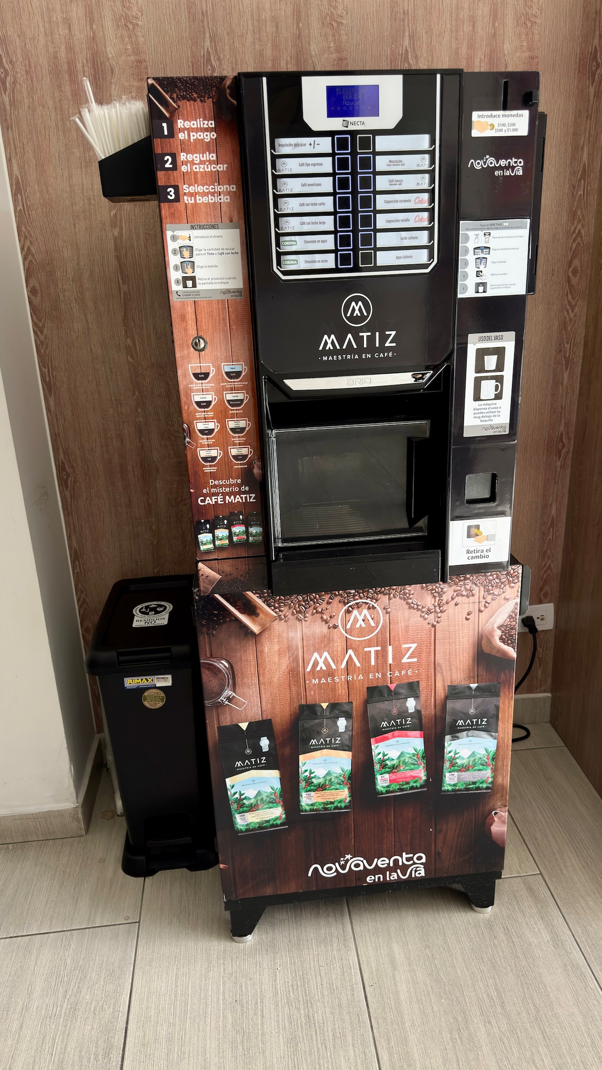 A beverage vending machine is displayed in a modern setting, featuring an array of drink options clearly labeled. The machine is positioned beside a small trash bin, and light-colored tiles create a neutral backdrop. The setting indicates accessibility for guests.