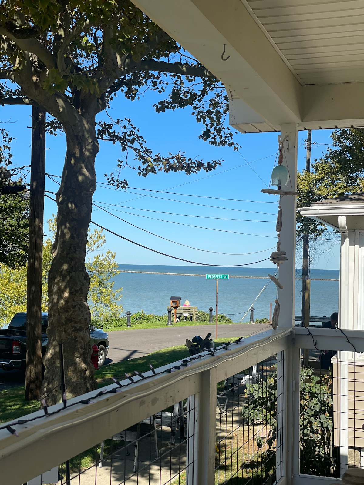 A serene view from the front porch showcases the calm lake, framed by trees and utility poles. The clear blue sky enhances the tranquil scene, while a glimpse of the street sign hints at the home's location. The railing features decorative elements, adding charm to the setting.