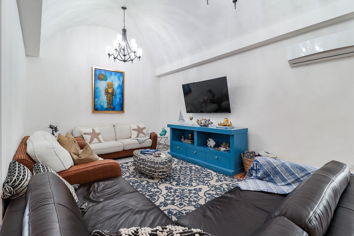 A spacious living area is presented, featuring a large sectional sofa upholstered in dark fabric and a light-colored sofa adorned with star motifs. A vibrant blue entertainment unit holds a flat-screen TV, while a decorative round coffee table sits on a patterned area rug.