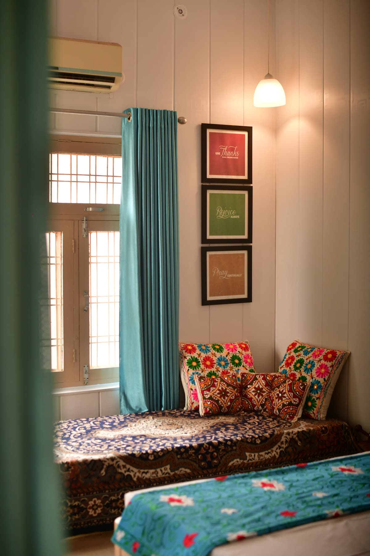 A cozy corner of the bedroom features a daybed adorned with colorful patterned cushions and a decorative throw. Curtains in a calming shade frame the window, while framed art pieces are arranged neatly on the wall above, adding a touch of character to the space.