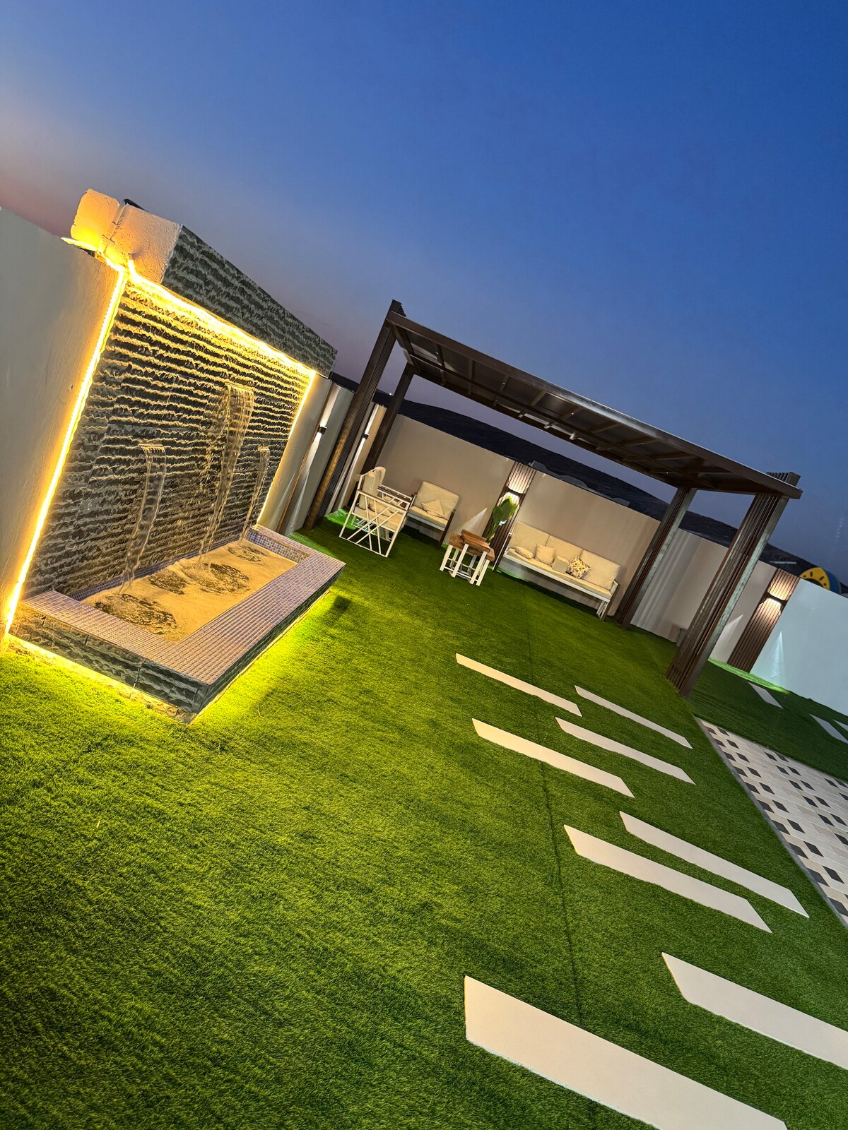 An outdoor seating area is showcased on a lush green lawn, illuminated by soft lighting. A wooden pergola provides shade over comfortable seating arrangements. A decorative water feature creates a calming atmosphere with gentle flowing water, complementing the tranquil space.