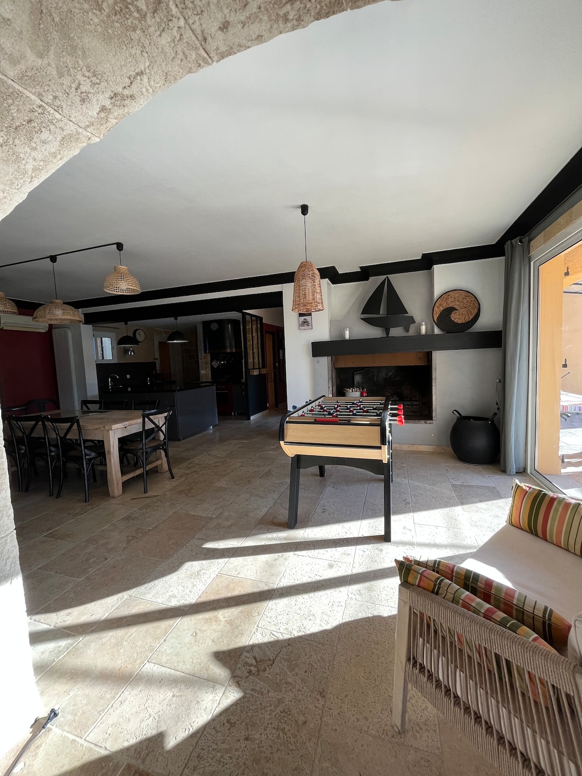 An open living area is highlighted by a foosball table near a stone fireplace. Soft lighting from woven pendant lamps creates a warm atmosphere. The space features a dining area with dark chairs and a kitchen visible in the background, while large glass doors lead outside.