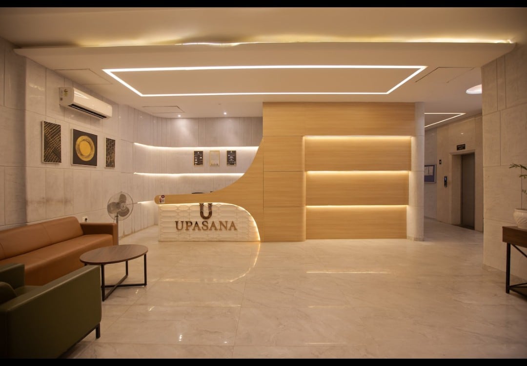 A modern lobby area features a sleek reception desk with the name 'UPASANA' prominently displayed. Soft lighting highlights the elegant design, while a combination of beige and wooden accents creates a serene ambiance. Comfortable seating options are available, alongside a small round table.