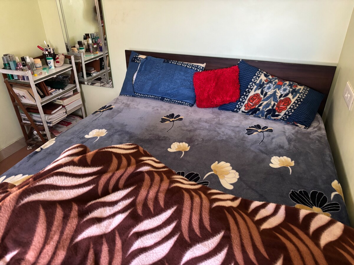 A cozy sleeping area features a spacious bed topped with a soft, floral-patterned blanket. Colorful cushions in blue and red add contrast against the neutral tones of the bedding. A mirrored dresser is visible in the background, showcasing neatly organized personal items.