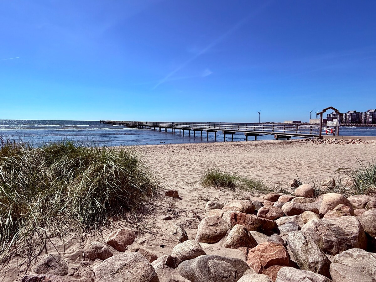 A sandy beach stretches towards a wooden pier that extends into the calm sea. Gentle waves lap against the shore, with tall grass and smooth stones scattered along the beach. A clear blue sky with a few wispy clouds creates a serene atmosphere.